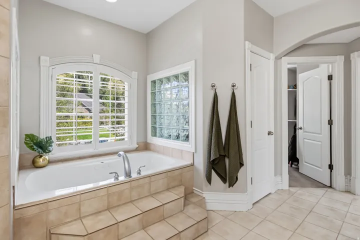Soaking tub with tiled surround, arched window with plantation shutters, glass block window, and tile flooring