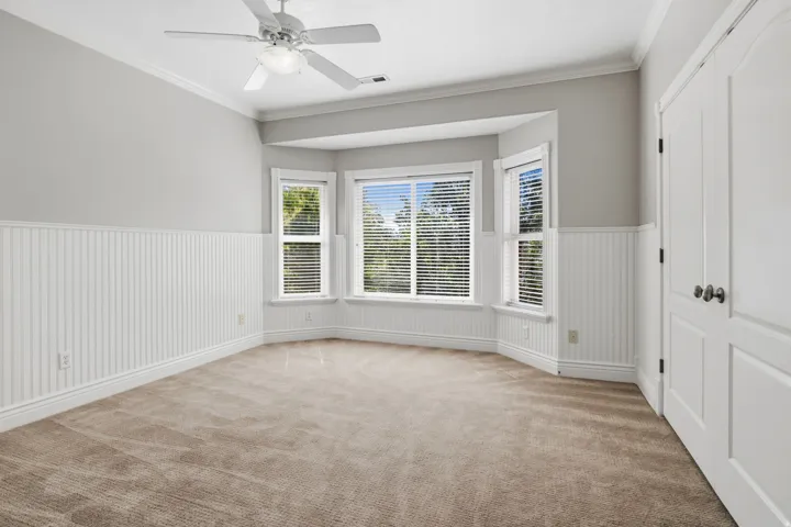 Bay window with blinds, ceiling fan with integrated lighting, crown molding, white beadboard wainscoting, and carpeted flooring