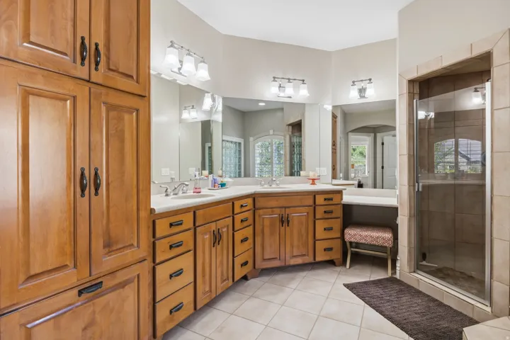 Dual vanity with integrated sinks, extensive wood cabinetry, tiled flooring, and a glass-enclosed shower