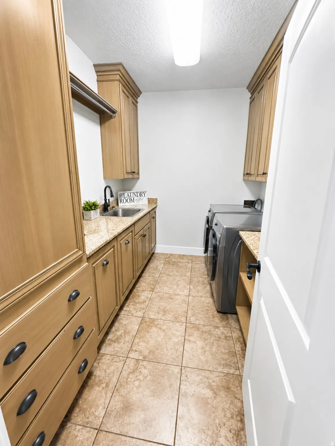 Functional laundry room featuring a utility sink, light wood-finish cabinetry, granite-look countertops, ceramic tile flooring, and integrated shelving