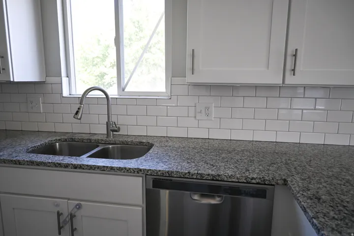 White shaker cabinetry, stainless steel double basin sink, stainless steel dishwasher, speckled countertop, and white subway tile backsplash