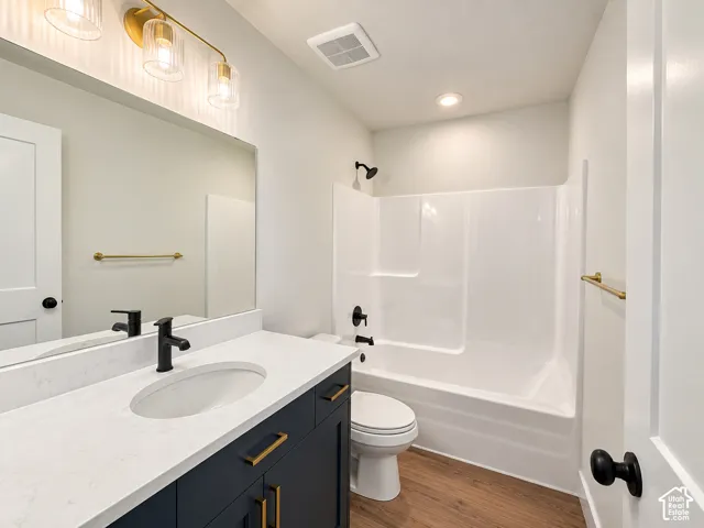 Vanity with white countertop and integrated sink, matte black faucet, gold-tone lighting fixture, white bathtub with matte black fixtures, and wood-finish flooring
