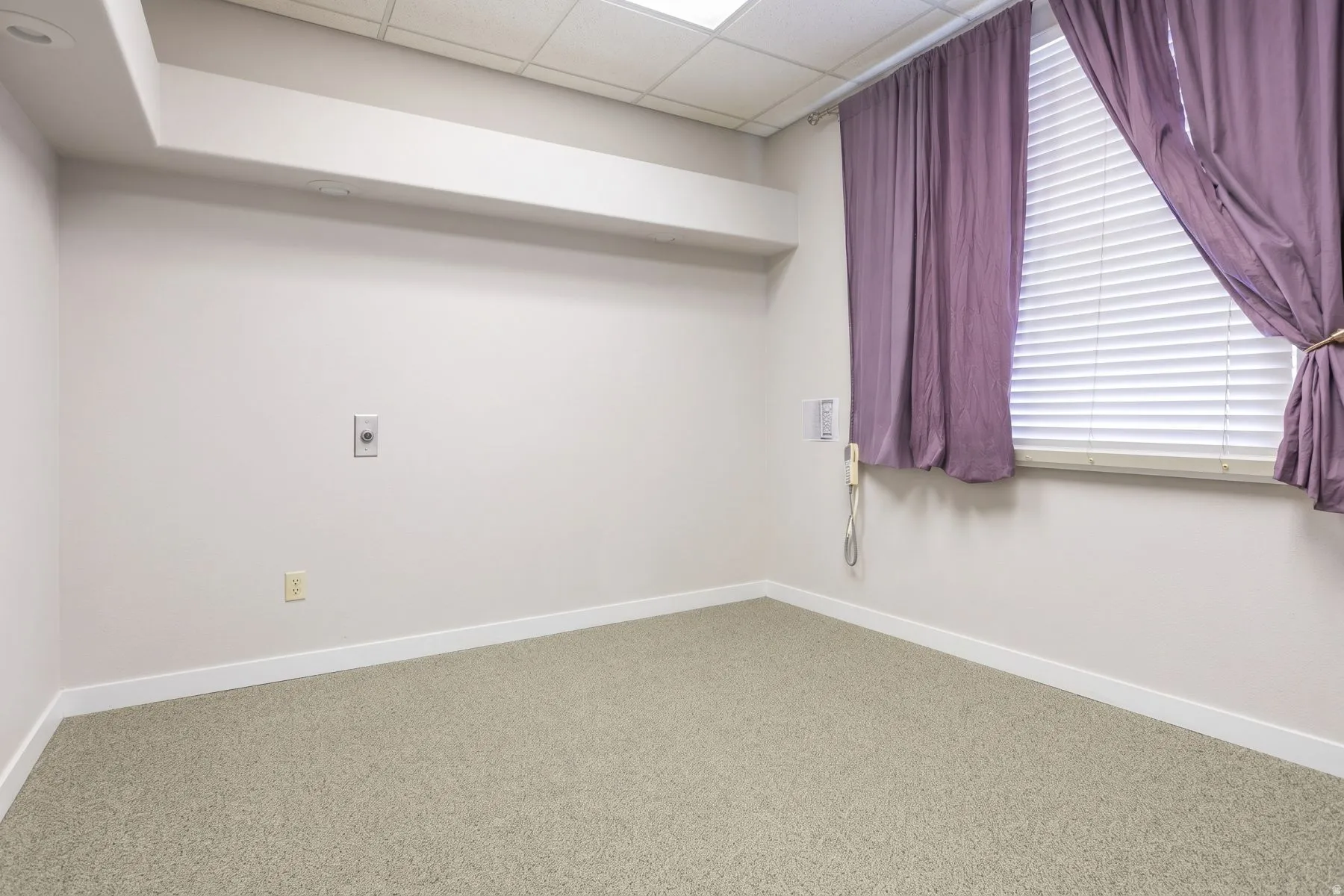 Textured carpet flooring, window with horizontal blinds, drop ceiling with recessed lighting, and a wall-mounted telephone jack