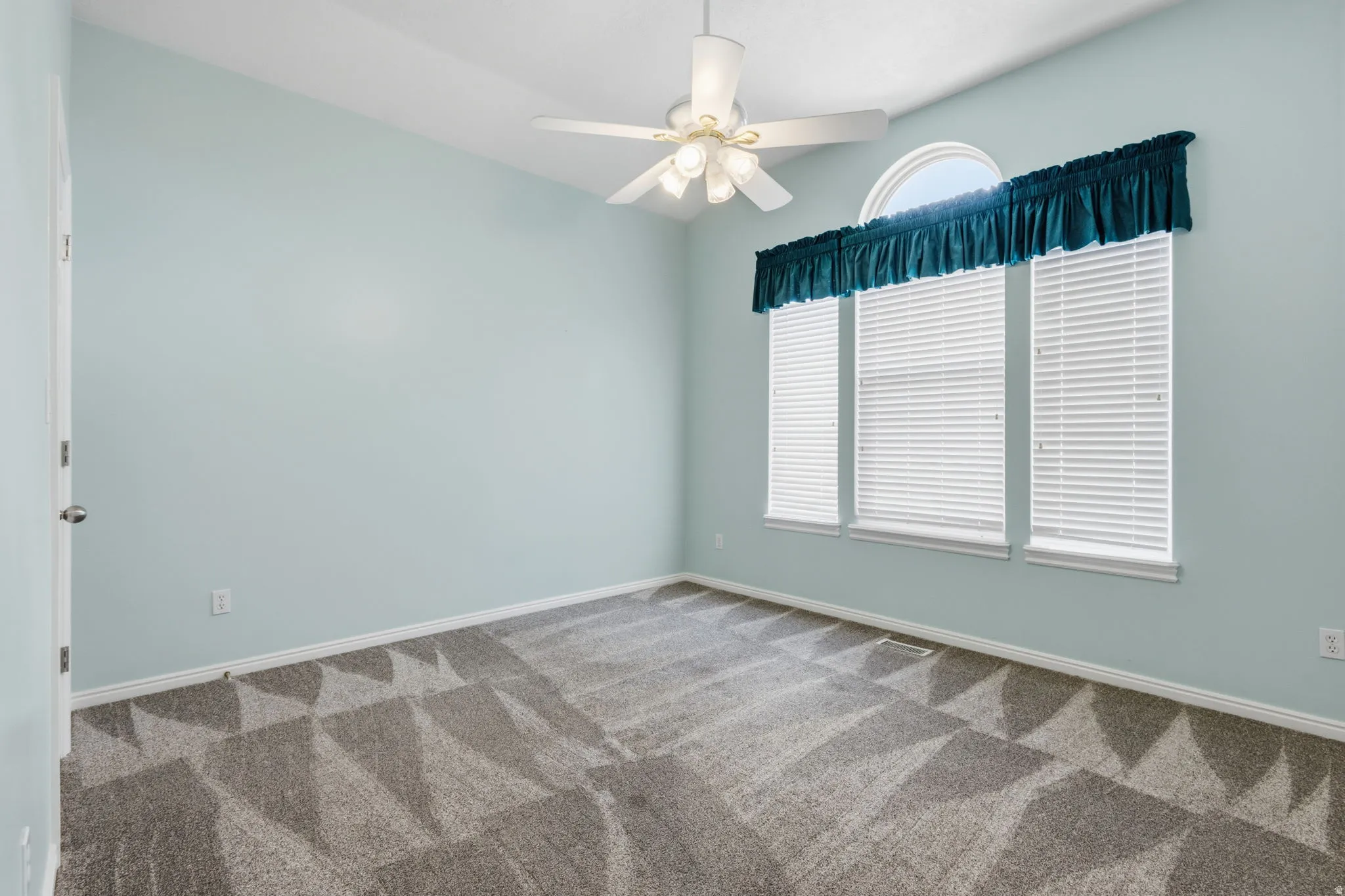 Vaulted ceiling, ceiling fan, three windows with blinds, arched transom window, and patterned carpeting