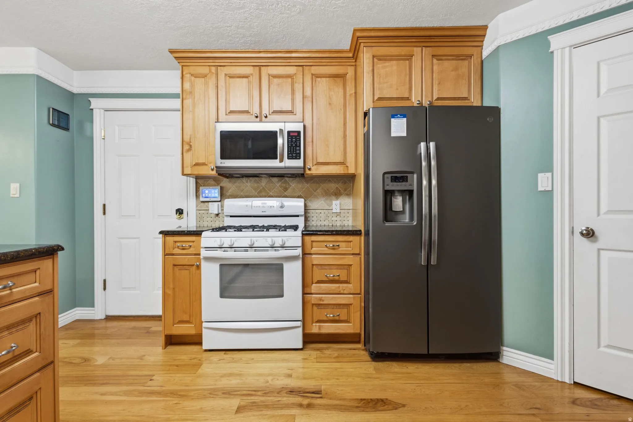 Kitchen featuring wood-finish flooring, natural wood cabinetry, dark countertops, a tile backsplash, and stainless steel appliances