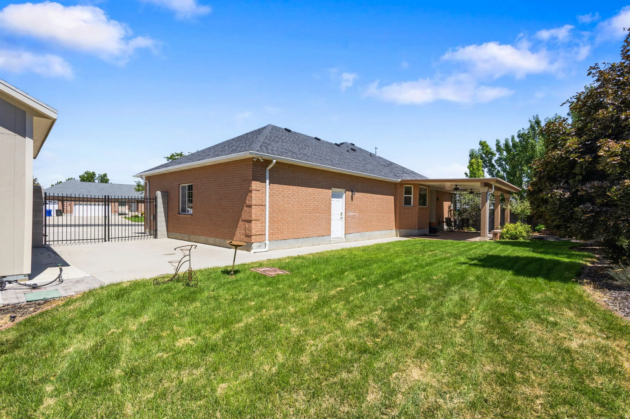 Expansive backyard featuring a manicured lawn, brick exterior, covered patio, and concrete driveway with metal gates