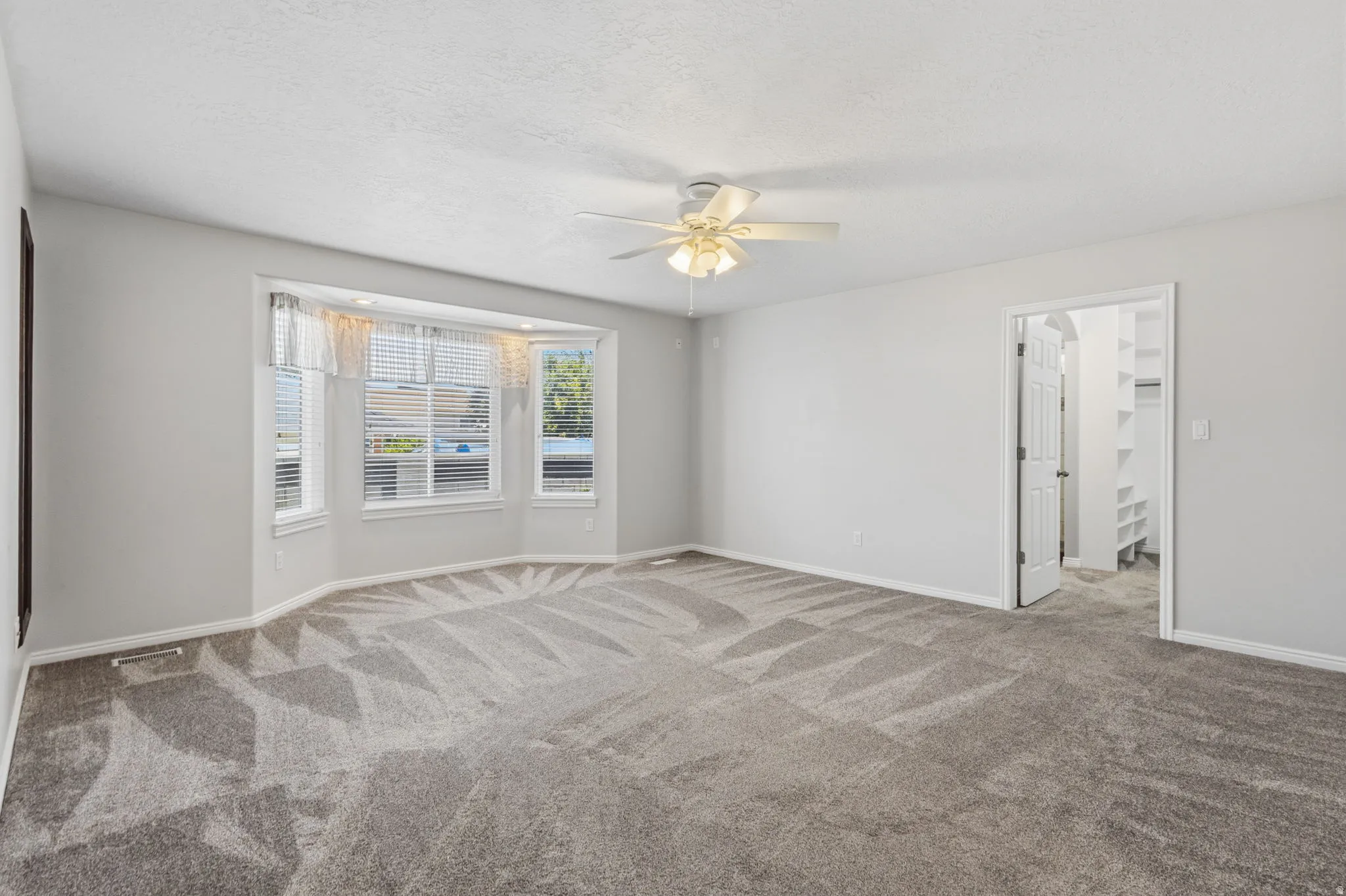 Bay window with blinds, ceiling fan, neutral wall paint, gray patterned carpet, and an open doorway to a closet with built-in shelving