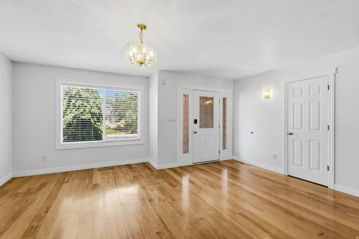 Hardwood flooring, white painted walls, large window with blinds, front entry door with decorative glass, and a brass-finish chandelier