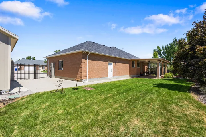 Expansive backyard featuring a manicured lawn, brick exterior, covered patio, and concrete driveway with metal gates