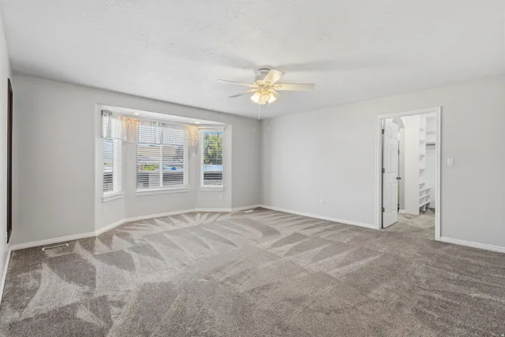 Bay window with blinds, ceiling fan, neutral wall paint, gray patterned carpet, and an open doorway to a closet with built-in shelving