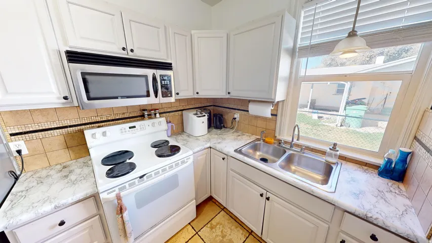 White cabinetry with dark hardware, stainless steel microwave, tile backsplash, laminate countertops, and a double basin stainless steel sink