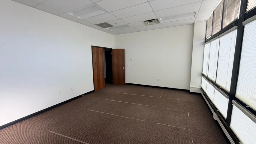 Carpeted room featuring a large window wall with horizontal blinds, wood-finish doors, and a dropped ceiling with recessed lighting