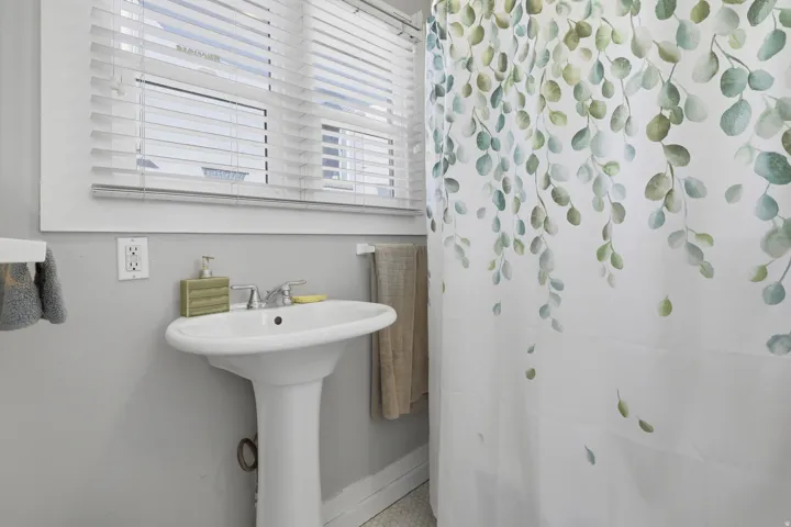 Pedestal sink with chrome faucet, window with white blinds, and light gray wall finish
