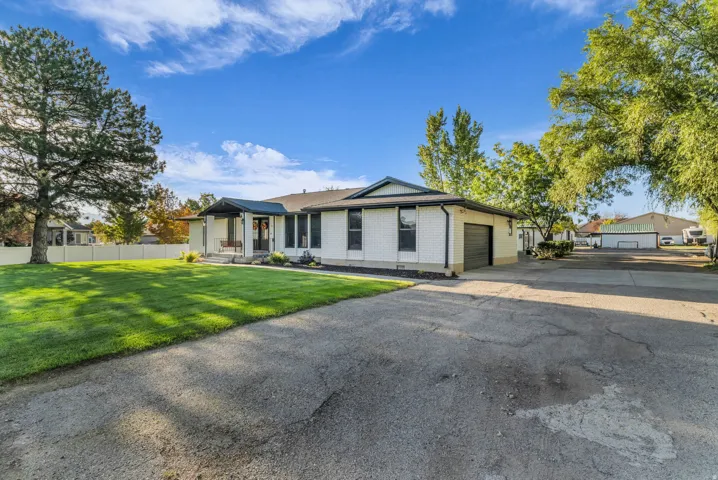 White brick exterior with dark trim accents, attached garage, expansive paved driveway, and mature landscaping