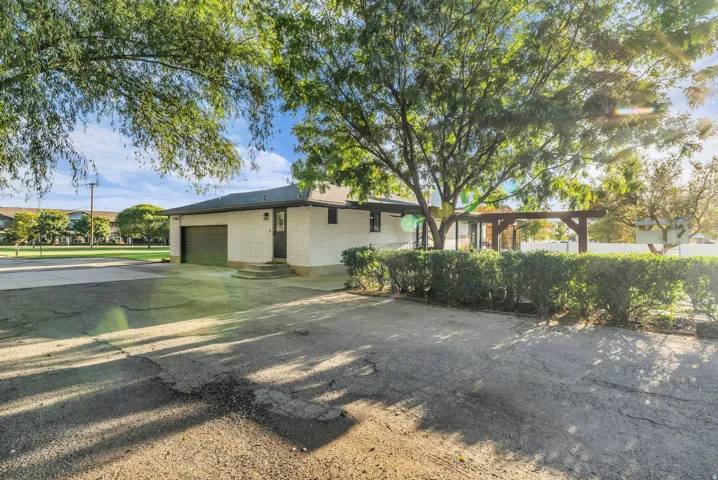 White exterior with attached garage, mature trees, wood pergola, and trimmed shrubbery