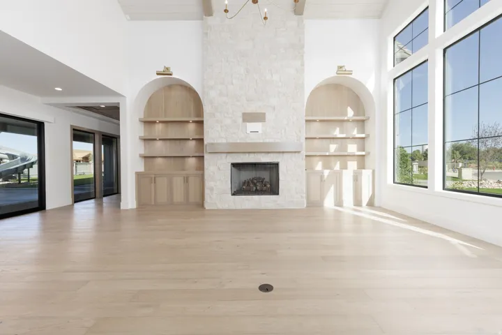 Light wood-finish flooring, stone-clad fireplace with mantel, built-in arched shelving with cabinetry, and expansive multi-pane windows