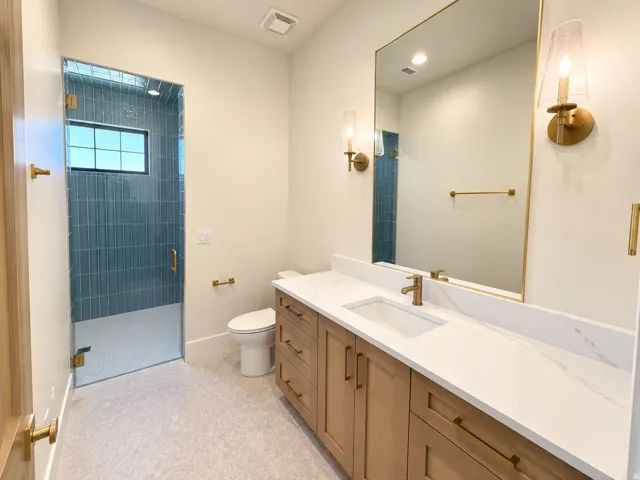 Glass-enclosed shower with vertical blue tile, light wood-finish vanity with white countertop, rectangular undermount sink, and wall-mounted sconces