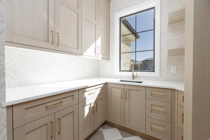 Built-in cabinetry with brass hardware, undermount sink with brass faucet, white countertop, patterned wall covering, and a multi-pane window