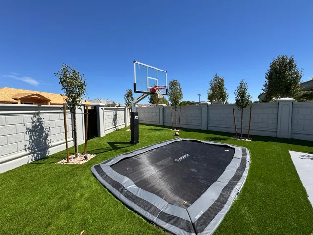 Expansive turf backyard featuring a built-in trampoline, basketball hoop, mature trees, and a decorative block wall