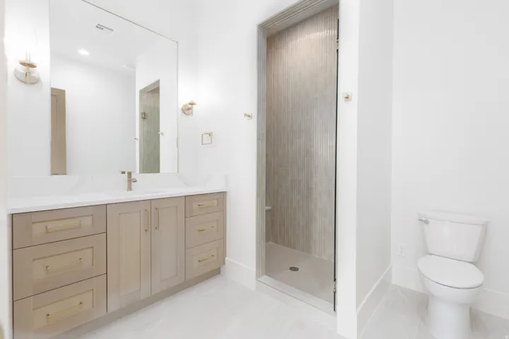 Bathroom featuring a double vanity with wood-finish cabinetry, a large mirror, wall-mounted sconces, a walk-in shower with vertical tile detailing, and light-toned floor tiling