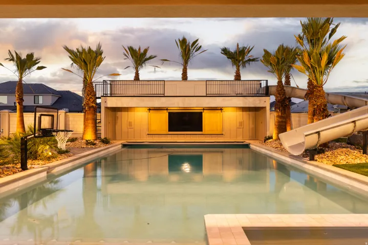 Expansive backyard featuring a large swimming pool, built-in water slide, upper-level balcony, ground-level outdoor entertainment area, and mature palm trees