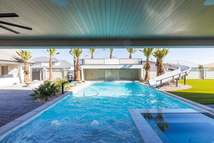 Expansive outdoor living area featuring a rectangular pool with an integrated spa, a water slide, a rock garden, and a covered patio with a ceiling fan