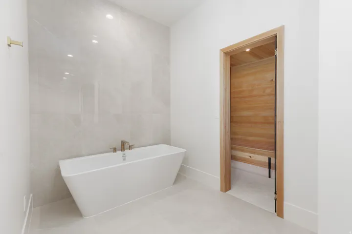 Freestanding soaking tub with a floor-mounted gold-tone faucet, large format tile walls, recessed lighting, and a wood-lined door frame