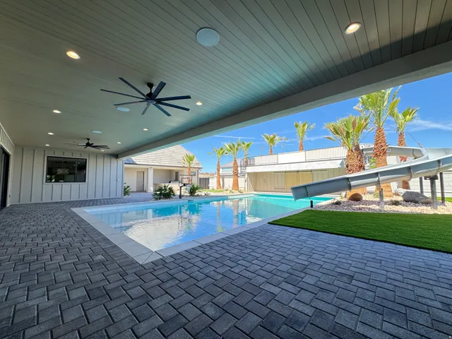 Expansive covered patio with recessed lighting and ceiling fans, opening onto a swimming pool with an integrated slide and artificial turf area, all surrounded by paver flooring