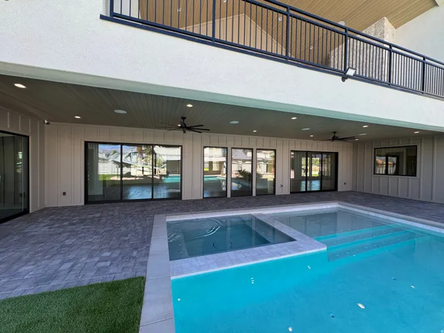 Expansive paved patio with an integrated pool and raised spa, featuring multiple sliding glass doors, built-in ceiling lighting, and a second-story balcony with dark metal railings