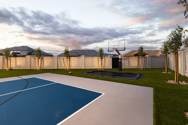 Expansive backyard featuring a dedicated sports court, artificial turf landscaping, a built-in trampoline, mature young trees, and a substantial privacy wall