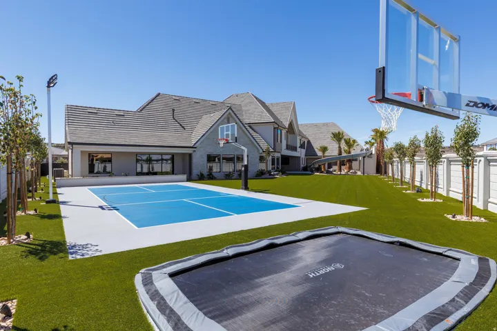 Expansive artificial turf backyard featuring a full-size sports court with a basketball hoop, mature trees, and a perimeter fence
