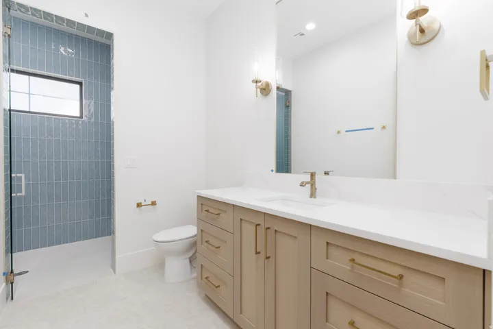 Glass-enclosed shower with blue vertical tile, single vanity with light wood cabinetry, white countertop, and gold-finish fixtures