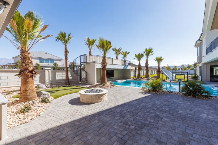 Expansive paver patio with a built-in fire pit, swimming pool with a slide, putting green, and mature palm trees