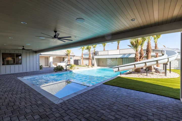 Expansive outdoor living area with a covered patio, dual ceiling fans, a swimming pool with an integrated spa, and a water slide feature