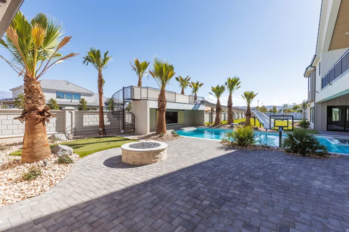 Expansive outdoor living area featuring a paved patio, swimming pool with a slide, putting green, fire pit, and mature palm trees