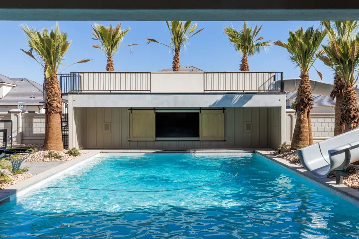 Pool with a built-in water slide, an integrated outdoor entertainment structure featuring a screen and barn door-style closures, and an upper-level balcony with metal railing
