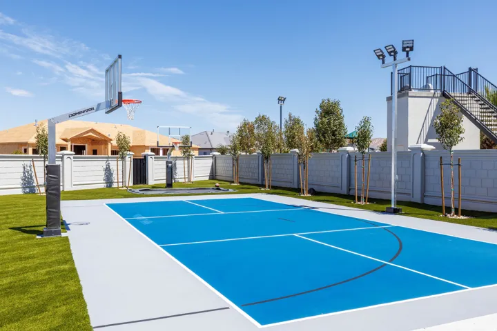 Outdoor sports court with a blue playing surface, integrated basketball hoop, and perimeter lighting
