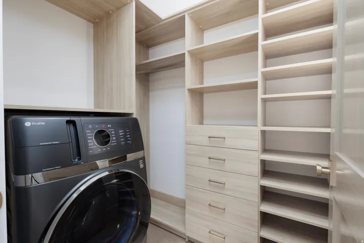 Integrated laundry appliance with built-in cabinetry, shelving, and drawers, featuring light wood-finish materials and brushed gold hardware
