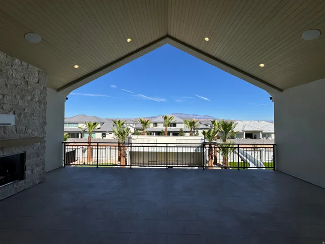 Expansive covered patio featuring a stone-clad fireplace with a wood mantel, recessed lighting, and a metal railing overlooking mature palm trees