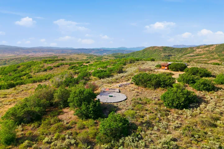 Expansive natural landscaping with a natural wood-sided structure, a round hardscape patio, and surrounding shrubbery