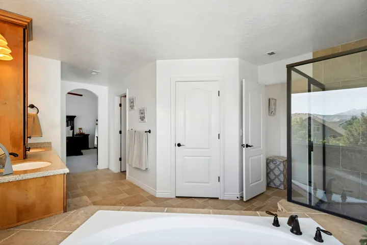 Large soaking tub with dark bronze fixtures, glass-enclosed shower, light wood cabinetry, dual vanity with undermount sinks, and tile flooring