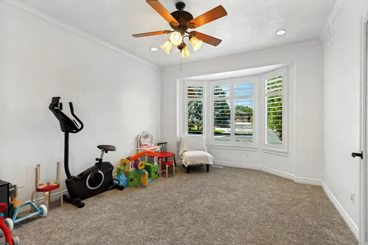 Bay window with plantation shutters, ceiling fan with integrated lighting, recessed lighting, crown molding, and carpeted flooring