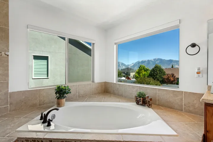 Built-in soaking tub with dark-bronze fixtures, surrounded by light-toned tiled surfaces, and large windows