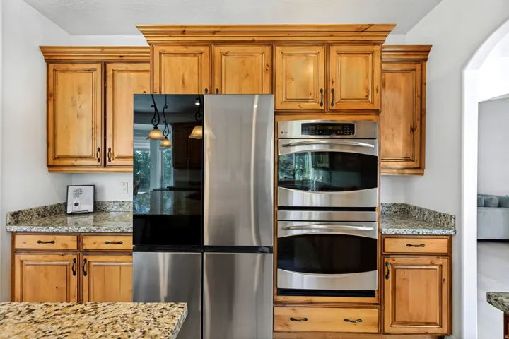Custom wood cabinetry with crown molding, granite-style countertops, stainless steel refrigerator, and built-in double ovens