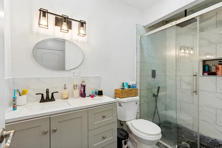 White quartz vanity countertop with integrated sink, contemporary vanity lighting, frameless oval mirror, floor-to-ceiling tiled shower with glass enclosure, and a built-in shower niche