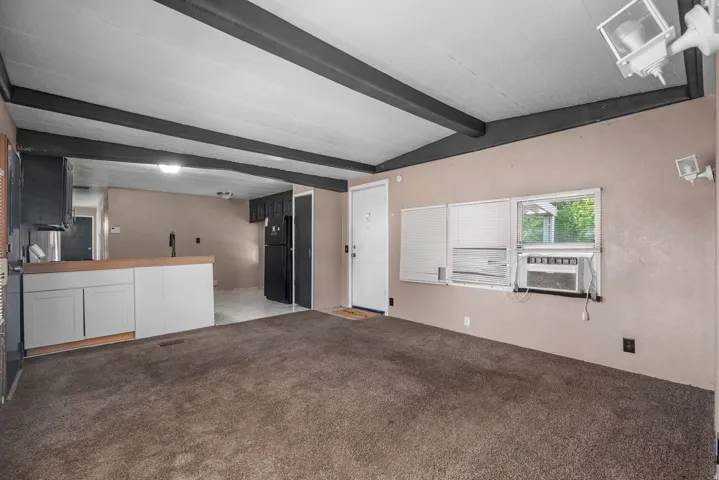 Open concept living area featuring exposed ceiling beams, a built-in kitchen counter with white cabinetry, dark wood-finish flooring in the kitchen, and brown carpet in the living space