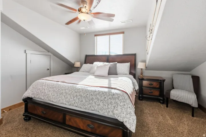 Sloped ceiling design, natural light from a window, overhead ceiling fan with lighting, and carpeted flooring