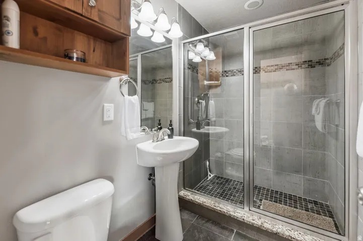 Pedestal sink, wood-finish wall cabinetry, glass-enclosed shower, tiled shower walls with accent mosaic, and dark tile flooring