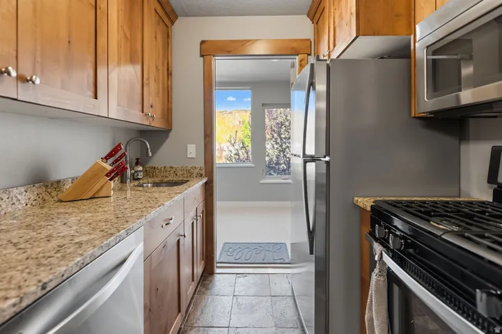 Galley kitchen with wood cabinetry, stone-finish countertops, stainless steel appliances, and a tile floor