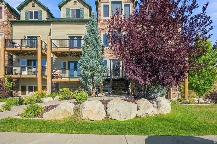 Multi-level structure with stone and siding exterior, featuring multiple balconies with black railings, a stone retaining wall, and mature landscaping with trees and shrubs